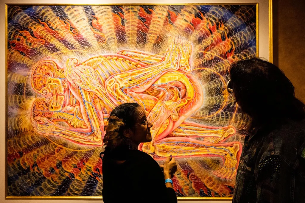 Two people viewing a psychedelic artwork by Alex Grey depicting entangled human forms with glowing energetic anatomy, often associated with DMT or spiritual visions